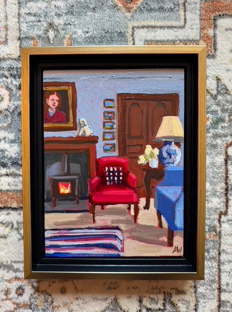 A scene of a living room with a red and blue armchair, a fireplace, and a portrait hanging above the fireplace. French doors are behind a side table and 5 pieces of art are hanging to the left of the doors. The fire is burning and there is a striped rug on the floor.