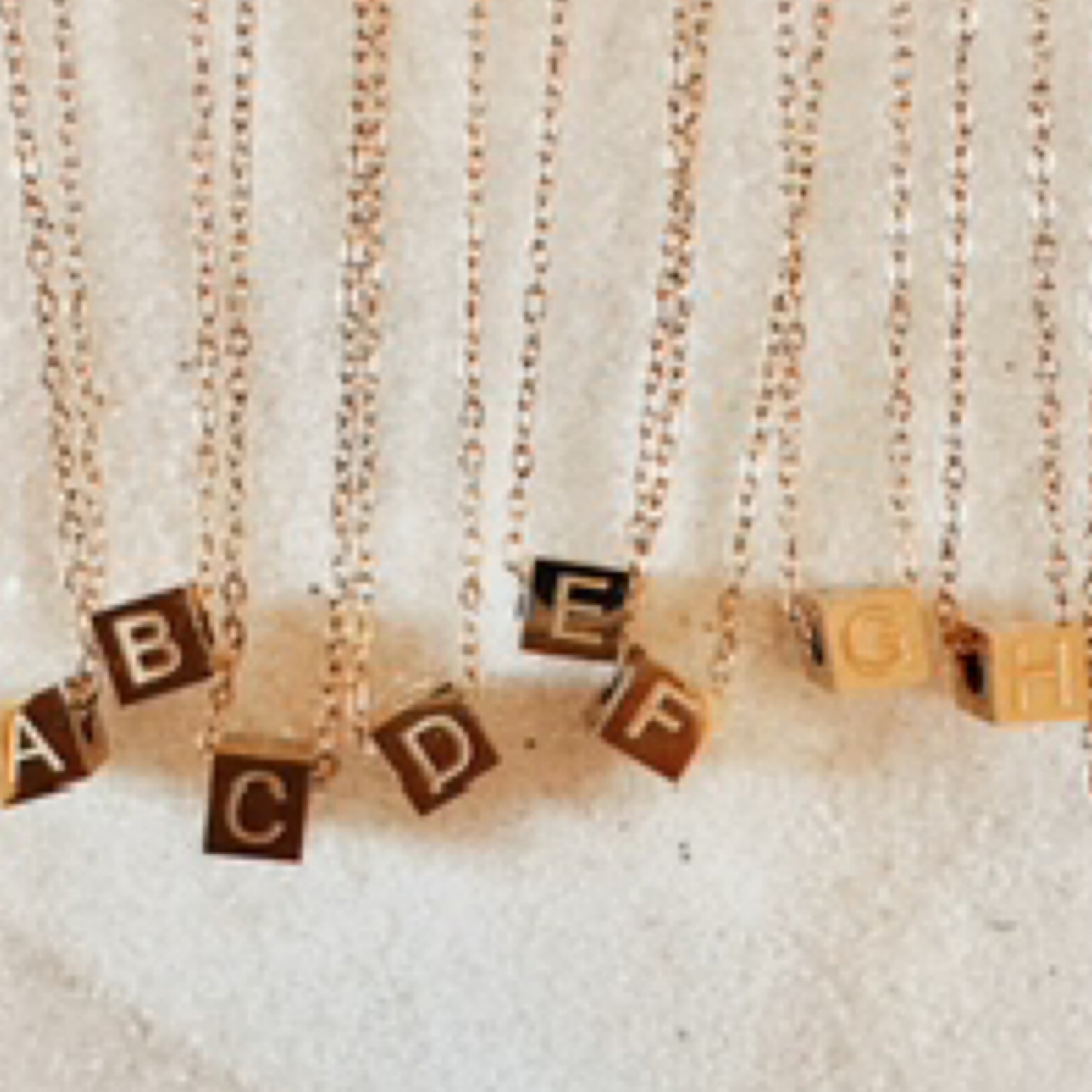 Range of gold  block charms with letters in graved on gold chains. Pictured against white sand.