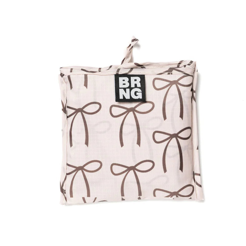 This image shows a compact, folded reusable shopping bag with a cream colored background and a pattern of brown bows. The bag is neatly packed into a square shape with a small fabric loop at the top and a "BRNG" logo label sewn on the front.