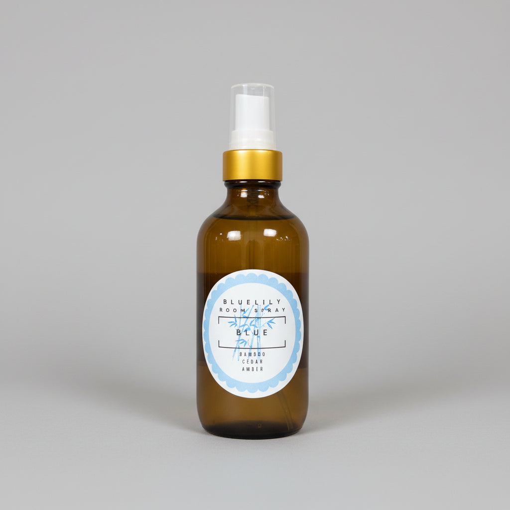An amber glass room spray bottle with a white spray top and a gold collar, featuring a circular white label with blue text that reads 'BLUELILY ROOM SPRAY' and 'BLUE'.