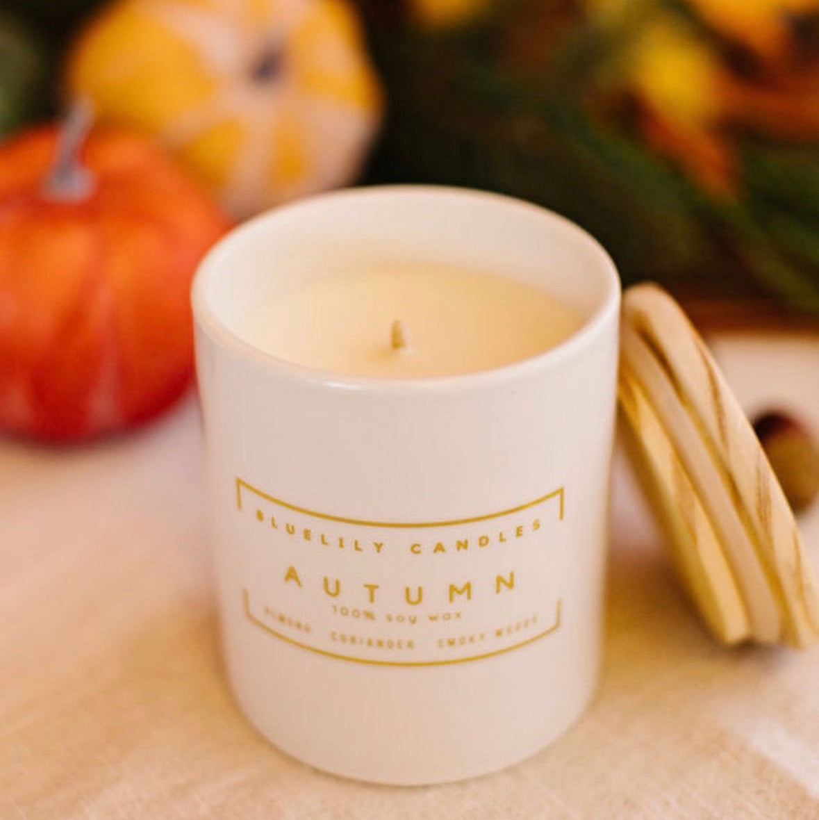 A white ceramic candle jar labeled 'Bluebilly Candles Autumn' with gold lettering, set on a light fabric surface with pumpkins and autumn foliage blurred in the background.