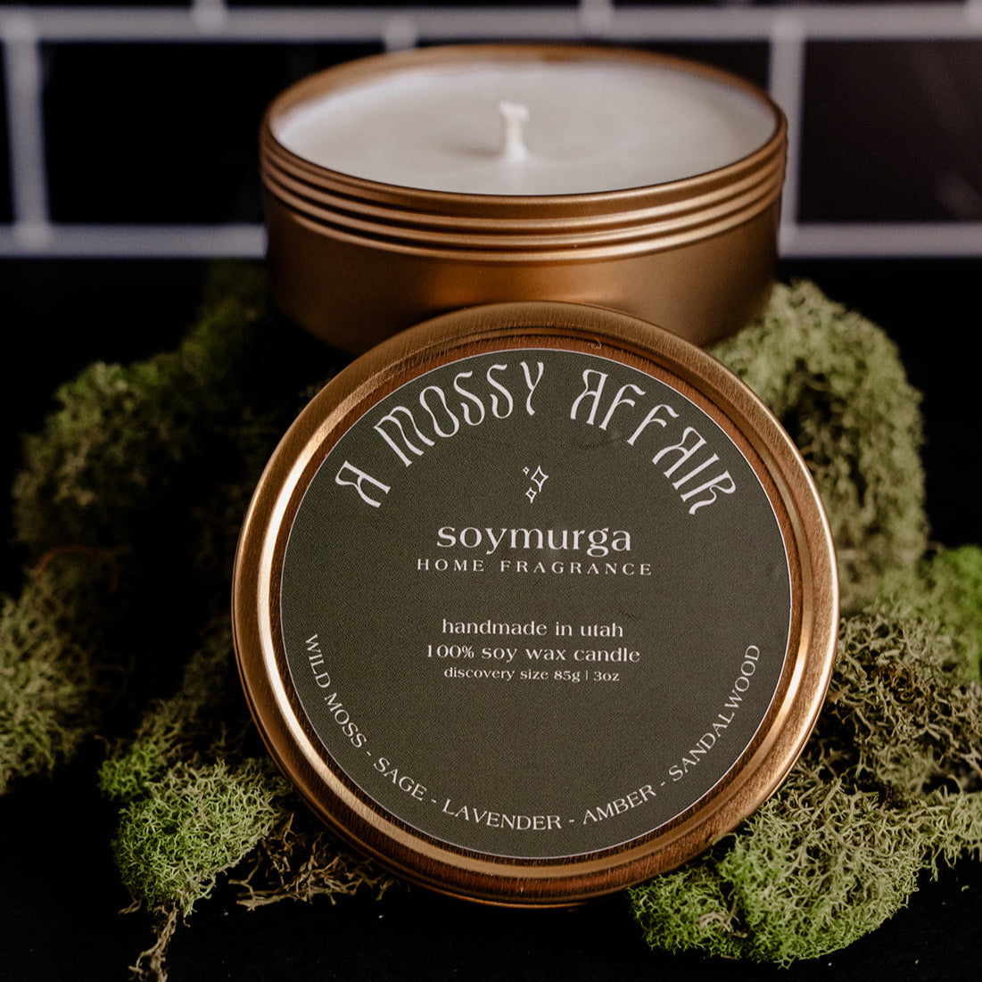 Small gold tin candle with a moss green label. Pictured atop a mound of moss with a black marble backs splash.