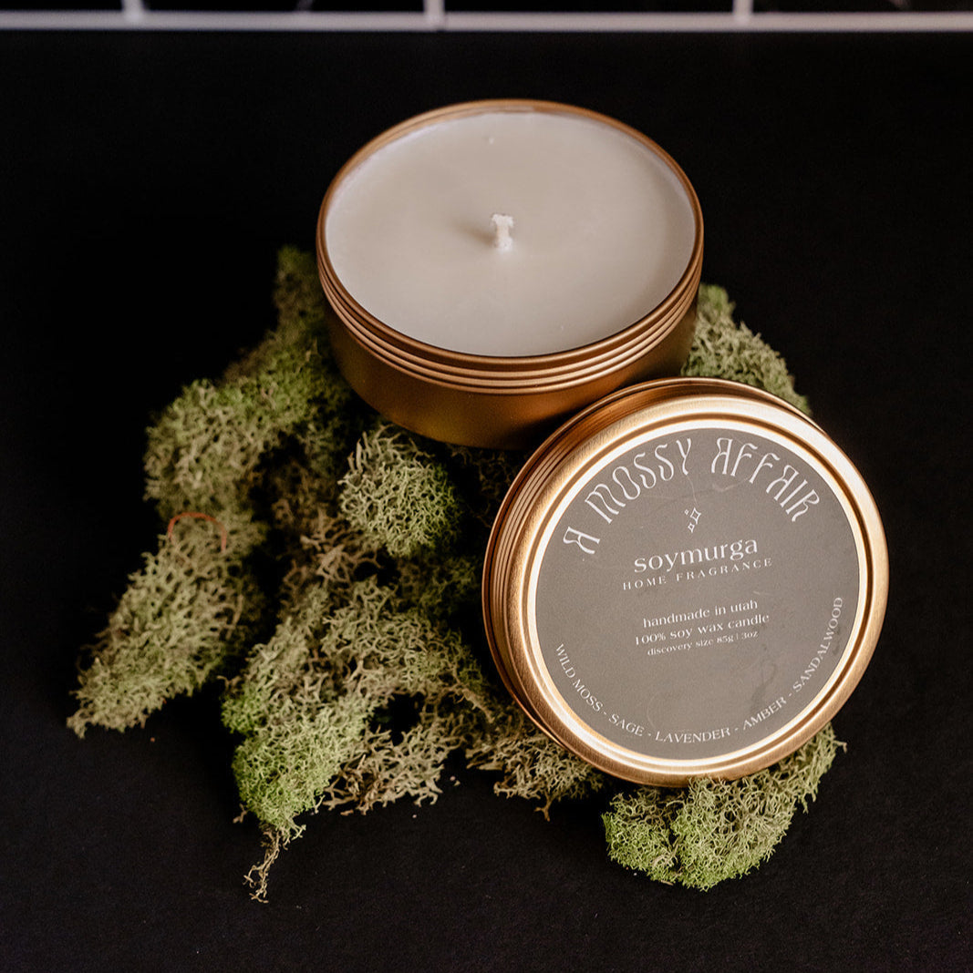 Small gold tin candle with a moss green label. Pictured atop a mound of moss with a black marble backs splash.