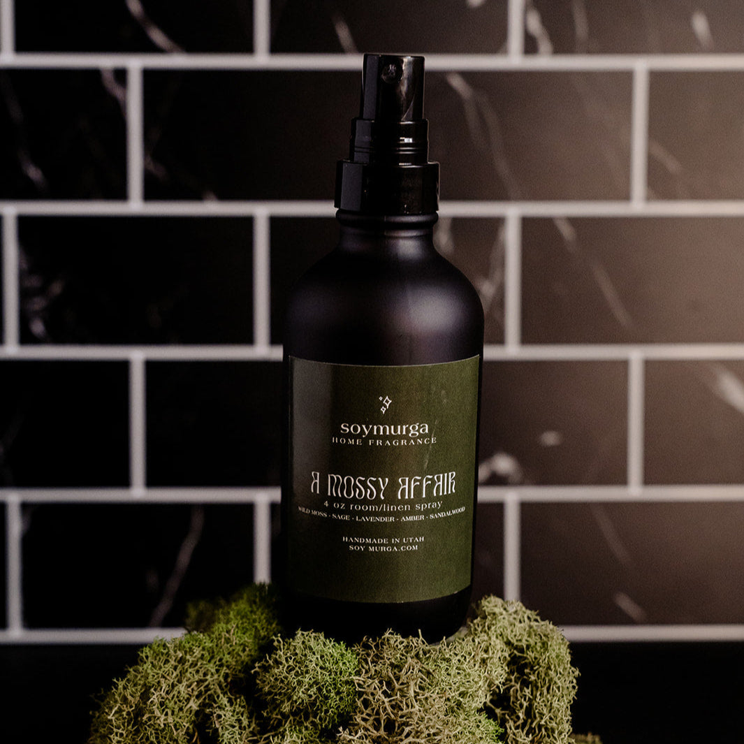 Black matte bottle with a moss green label. Pictured atop a mound off moss with a black marble back splash.