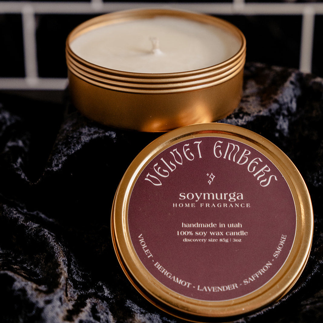 Small gold tin candle with a burgundy lable on lid which is sitting next to the candle on black velvet. Pictured against a black marble backsplash.