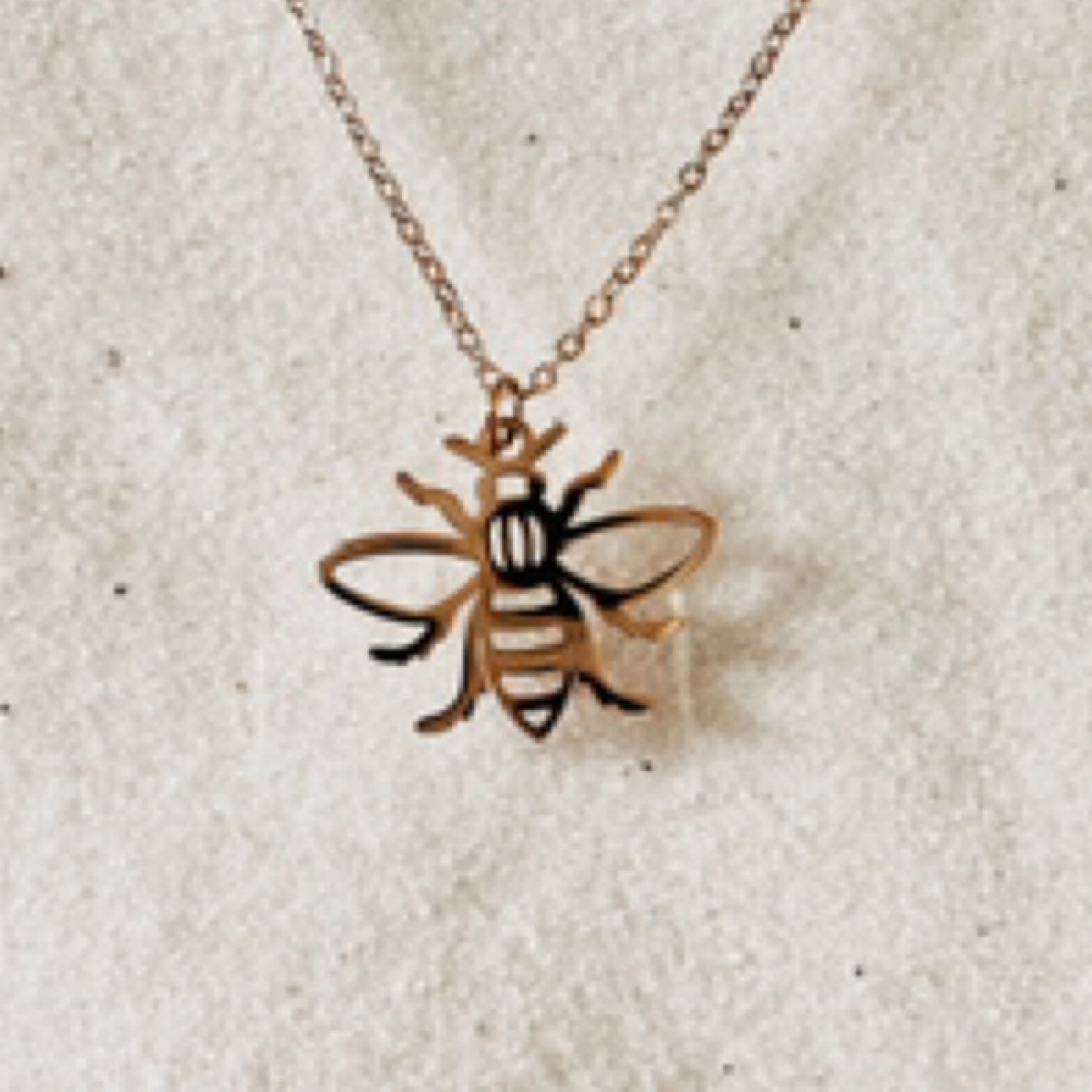 Delicate gold chain with a gold bee cutout charm. Pictured against white sand.