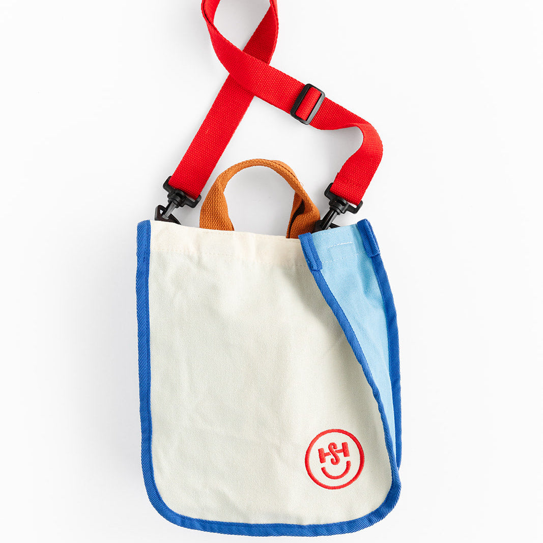 A cream-colored canvas tote bag with blue trim and orange handles features a red circular logo near the bottom corner. The bag includes a bright red adjustable shoulder strap with black clips against a white background.