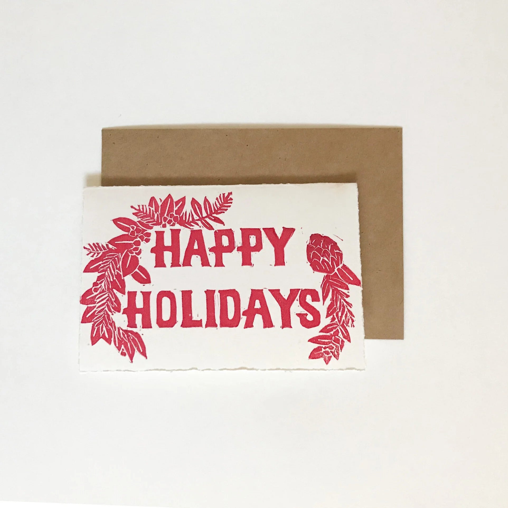 a relief linocut card with "HAPPY HOLIDAYS" written in the center with winter foliage framing two corners