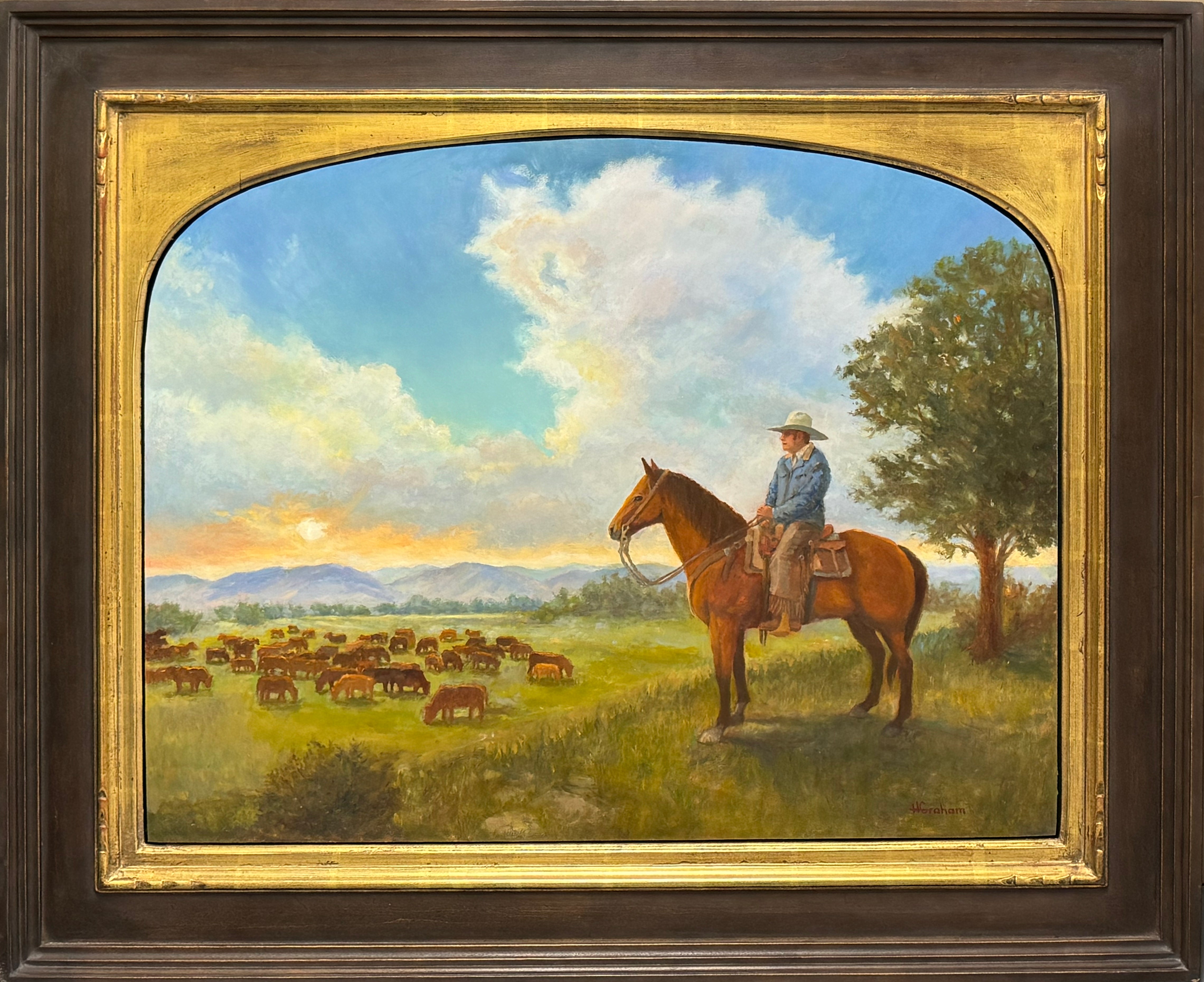Cowboy wearing a jean jacked and leather chaps with fringe and a beige cowboy hat sits atop his brown bay horse overlooking a heard of cows grazing a green pasture. The sky is bright blue with thin clouds and in the background is a big mountain range with the sun setting.