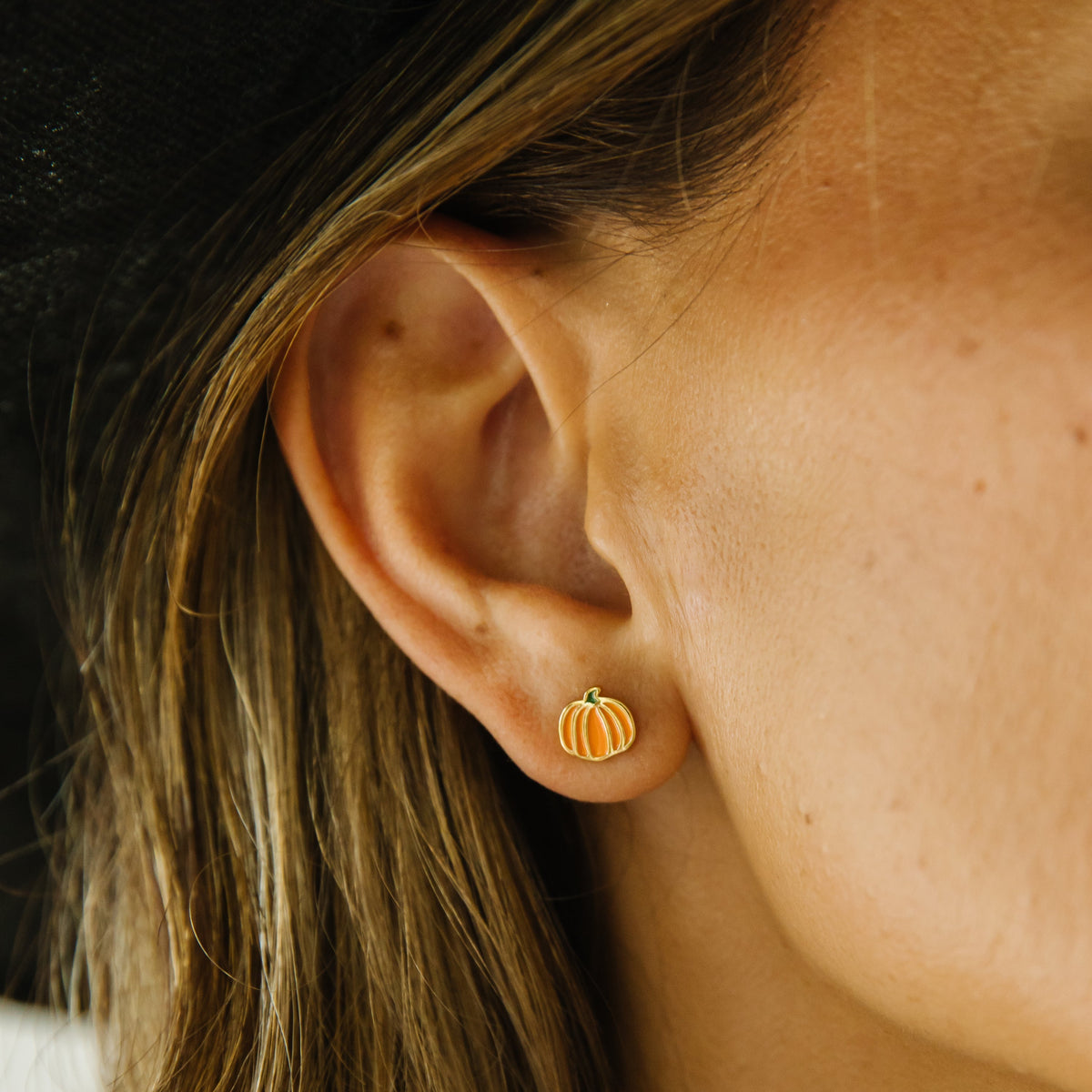 A close-up photo of a person wearing a small gold pumpkin-shaped stud earring, featuring a simple outlined pumpkin design with a stem.