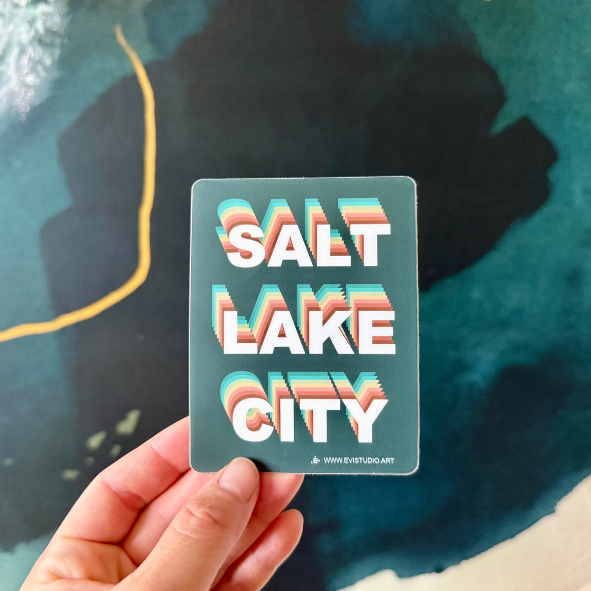 Dark green sticker with rainbow fade letters saying Salt Lake City. held in woman's hand.
