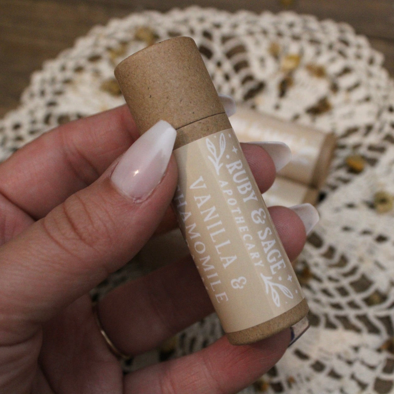 Brown paper lip balm tube with beige label with white font. Held in an hand with white nails, held above a doily and crushed chamomile on a wooden table.
