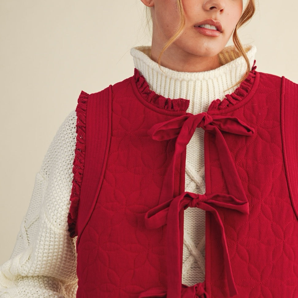 Elevate your layering game with this charming red floral quilted vest. Crafted from soft quilted fabric adorned with delicate floral patterns, it features a front-tie closure that lets you customize the fit to your comfort. The pretty lace trim adds a touch of feminine elegance to any outfit. Perfect for layering over your favorite tees or dresses, this versatile piece brings warmth and style to your everyday wardrobe.