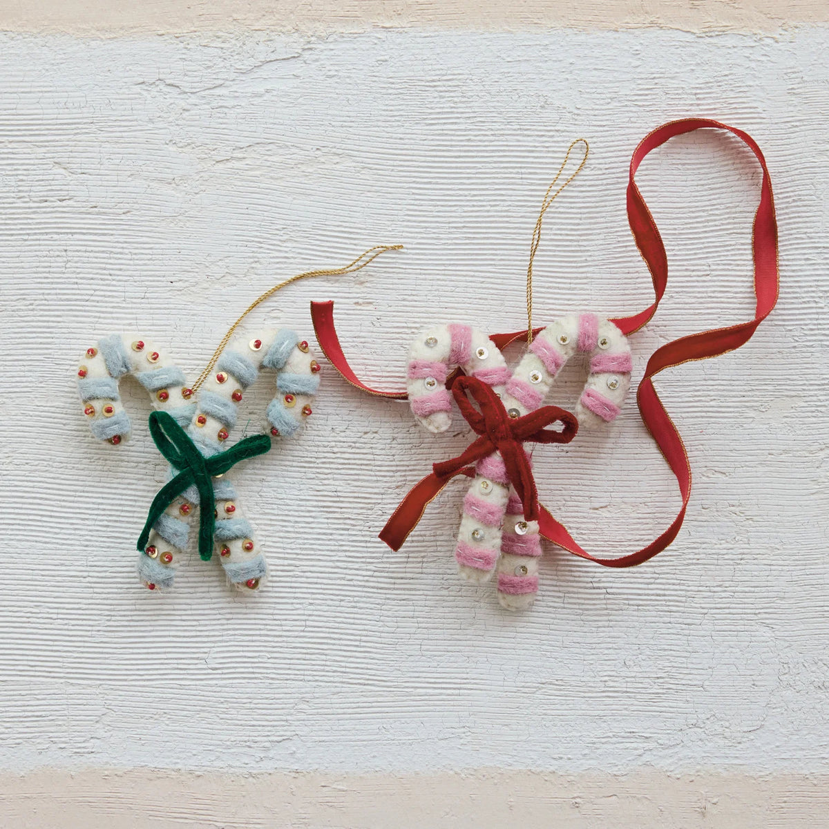 Two handmade felt holiday ornaments shaped like candy canes, made from white and pink striped fabric with small embroidered details, hanging from thin twine with red ribbons, displayed on a textured off-white background.