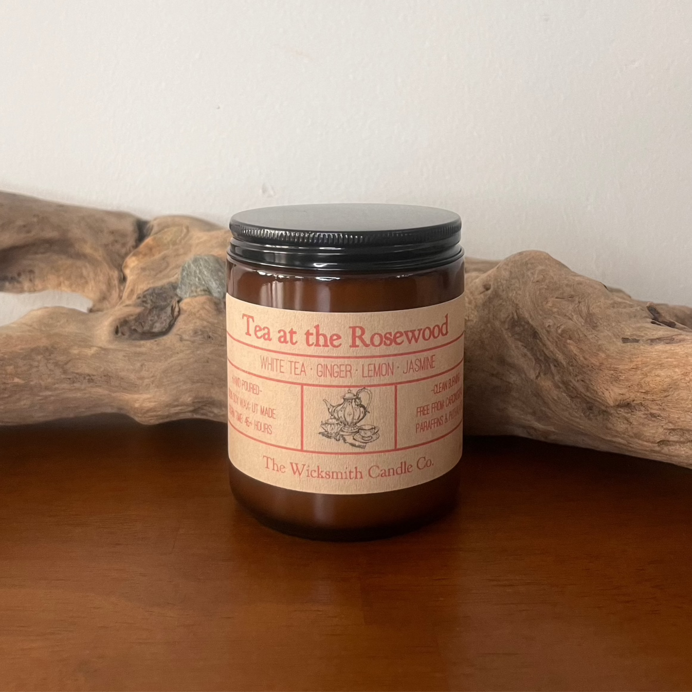 Amber glass candle jar labeled ‘Tea at the Rosewood’ by The Wicksmith Candle Co., featuring scent notes of white tea, ginger, lemon, and jasmine placed on a wooden surface with a piece of driftwood in the background