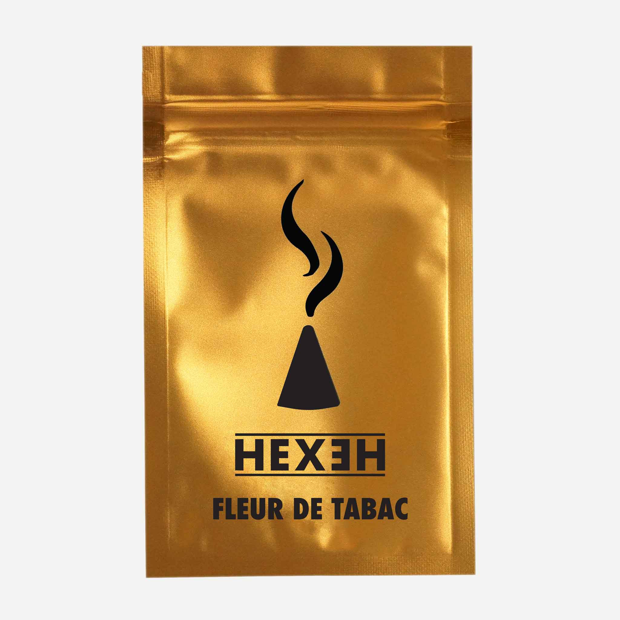 A shiny gold metallic pouch features a central black graphic of a stylized incense cone with rising smoke curls above it, accompanied by the brand name "HEXEH" in bold uppercase letters and the product name "FLEUR DE TABAC" below. The pouch is photographed straight-on against a plain light background, highlighting its reflective surface and clean, minimalist design.
