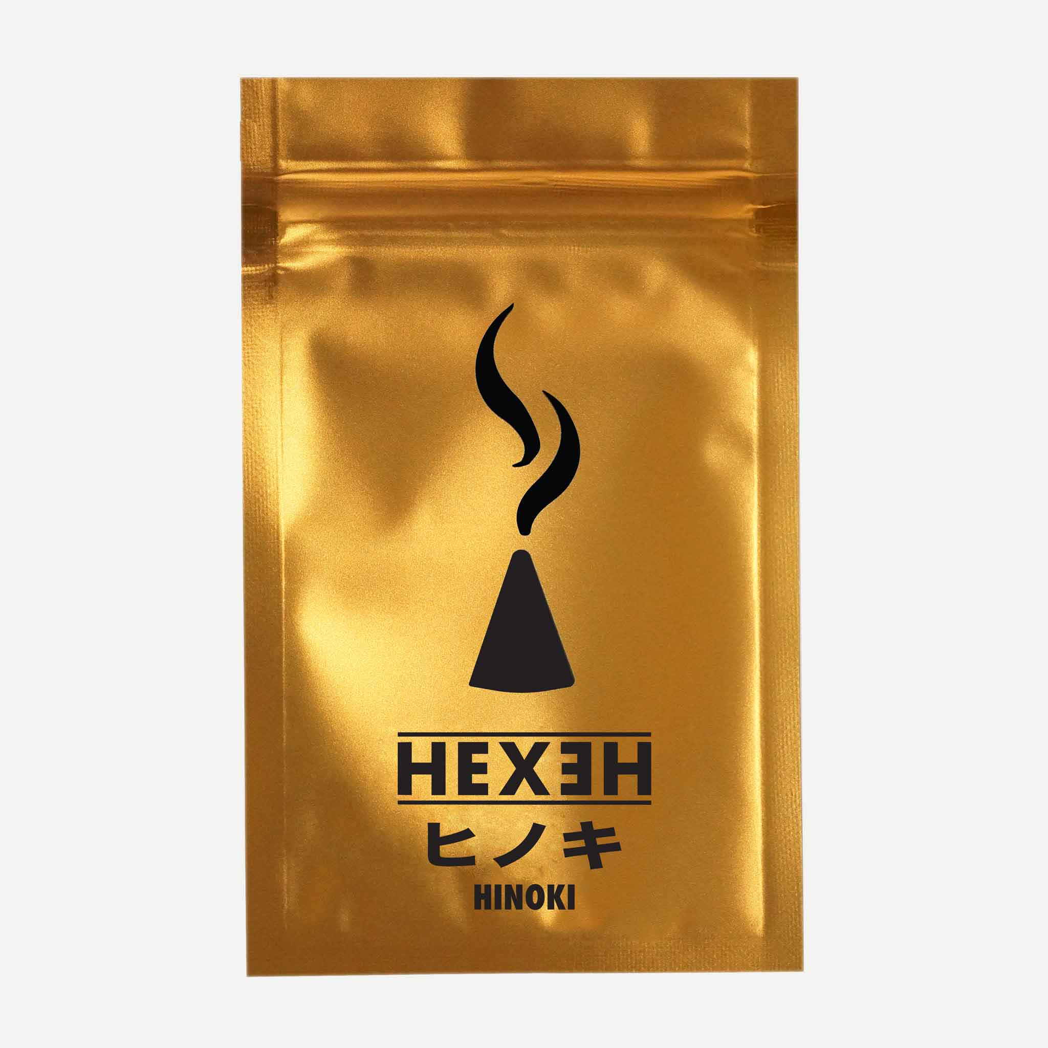 A shiny gold metallic pouch features a central black graphic of a stylized incense cone with rising smoke curls above it, accompanied by the brand name "HEXEH" in bold uppercase letters and the product name "HINOKI" below. The pouch is photographed straight-on against a plain light background, highlighting its reflective surface and clean, minimalist design.