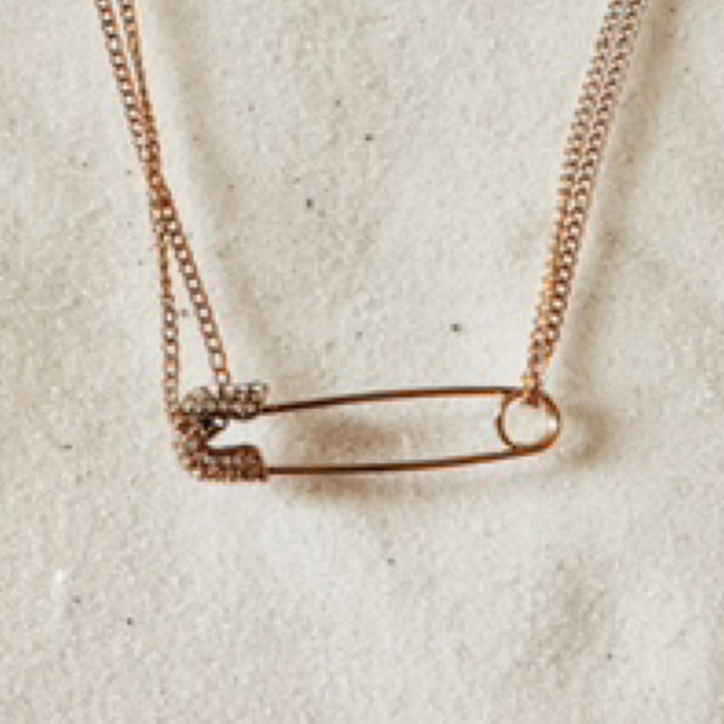 Gold double strand chain with a bedazzled sparkly safety pin charm hanging horizontally. Pictured against white sand.