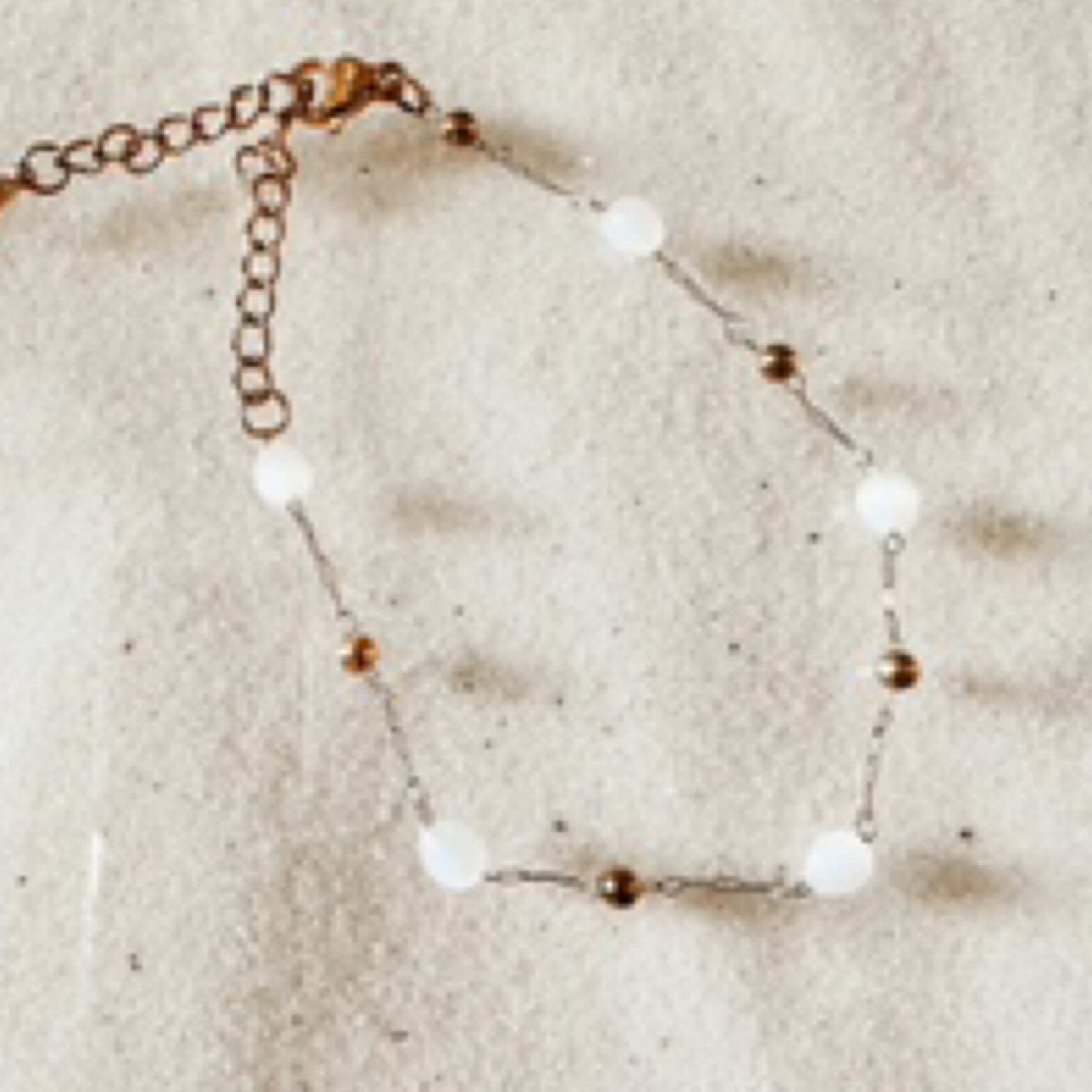 Delicate gold chain with small gold beads and moonstone beads spread out. Pictured on white sand.