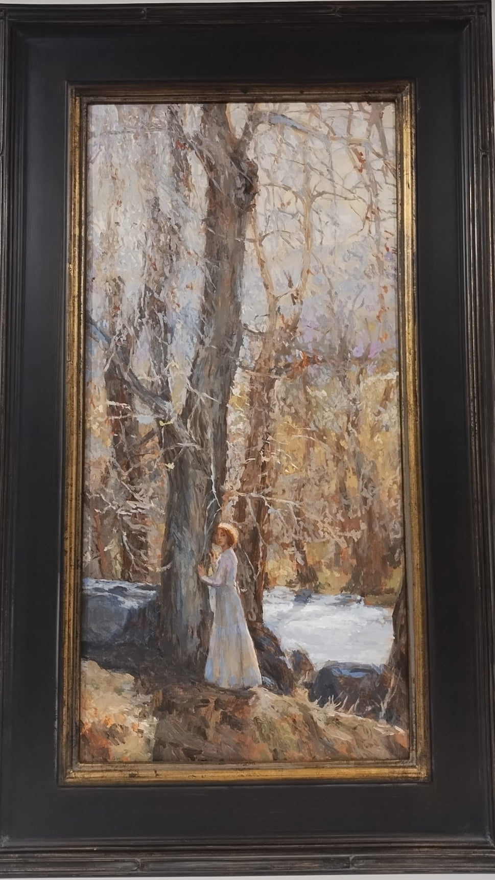 A distant view of a young woman standing next to a tree with her hands against the trunk. She has her head turned toward you and she has golden red hair in a bob. She is wearing a flowy white dress that goes down to her ankle. The tree is almost three times her height and the sun is shining from the background. There is some snow on the ground and a golden hue.