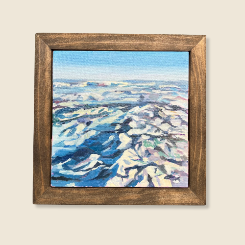 Size 6"x6". Oil on canvas. Framed in dark natural wood frame. Ariel view of mountains in snow, dark shadows contrast with white slopes, blue sky, layer of clouds