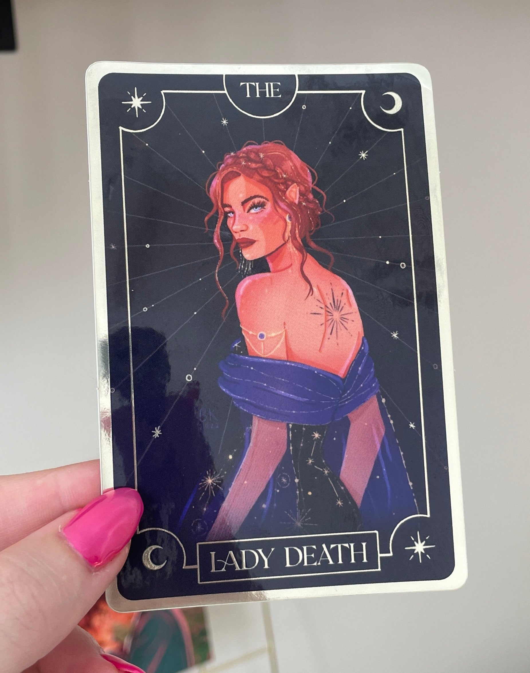 Nesta "The Lady Death" is on a sticker resembling a tarot card. Nesta has her brown hair braided and in an updo and she is lookng back over her shoulder at you. She has a tattoo of an exploding star in the center of her upper back and she is wearing a deep blue dress adorned with constellations.