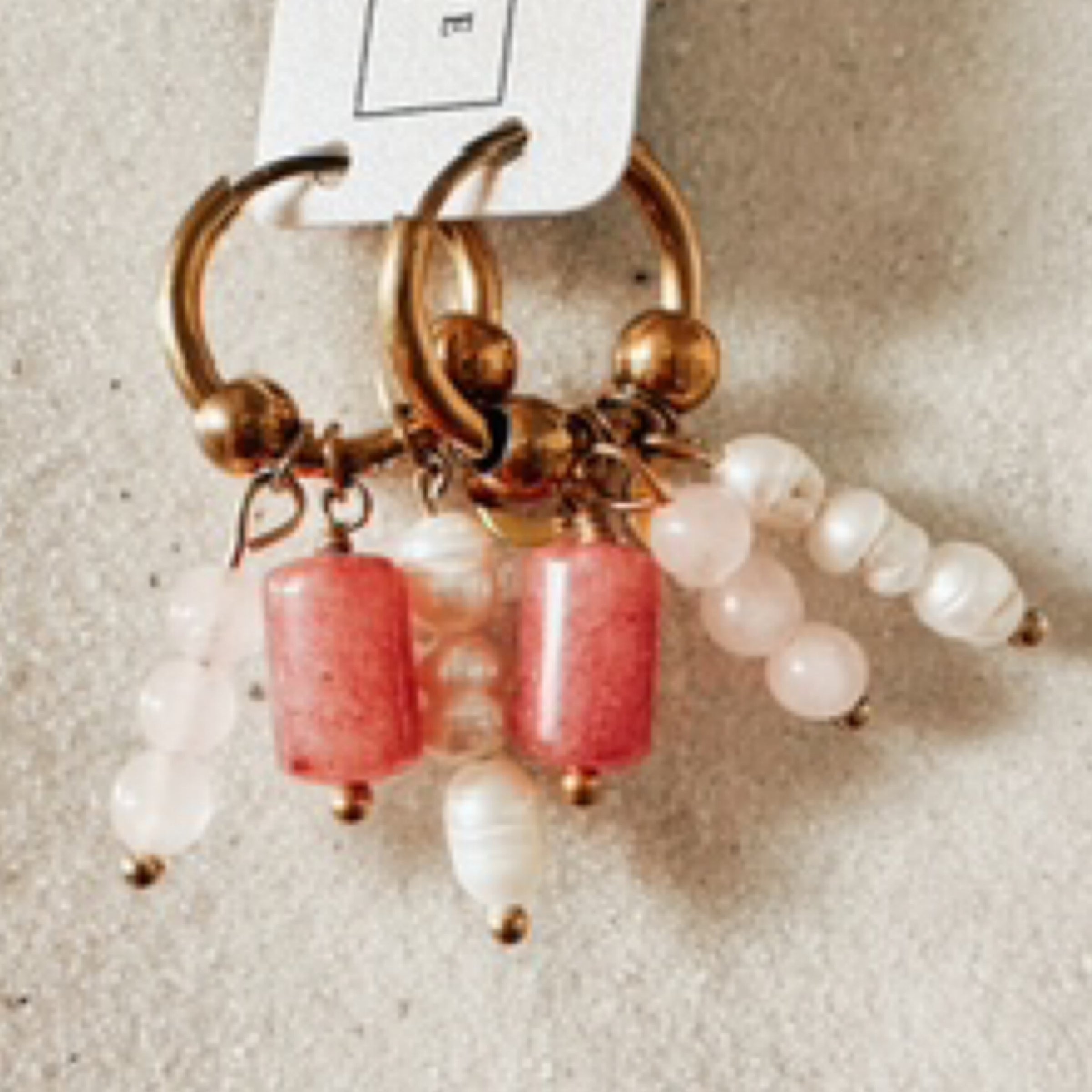 Gold hoops with multiple bead charms and a tiny gold crescent moon charm (1 pearl bead stack charm, 1 cylinder shaped pink stone charm, 1 light pink stone ball stack charm.) Pictured against white sand.