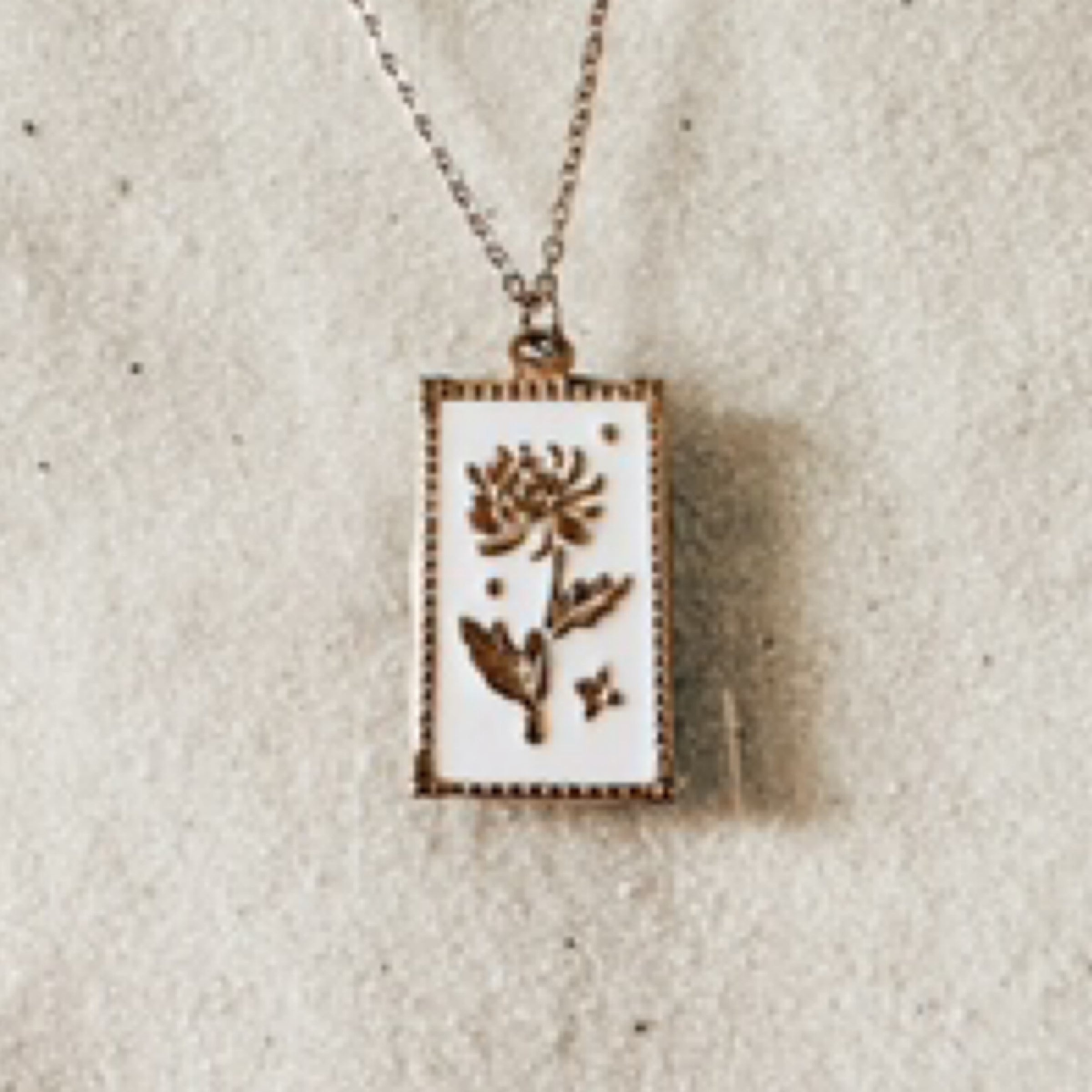 Delicate gold chain with a square white pendant stamped with a gold chrysanthemum. Pictured against white sand.