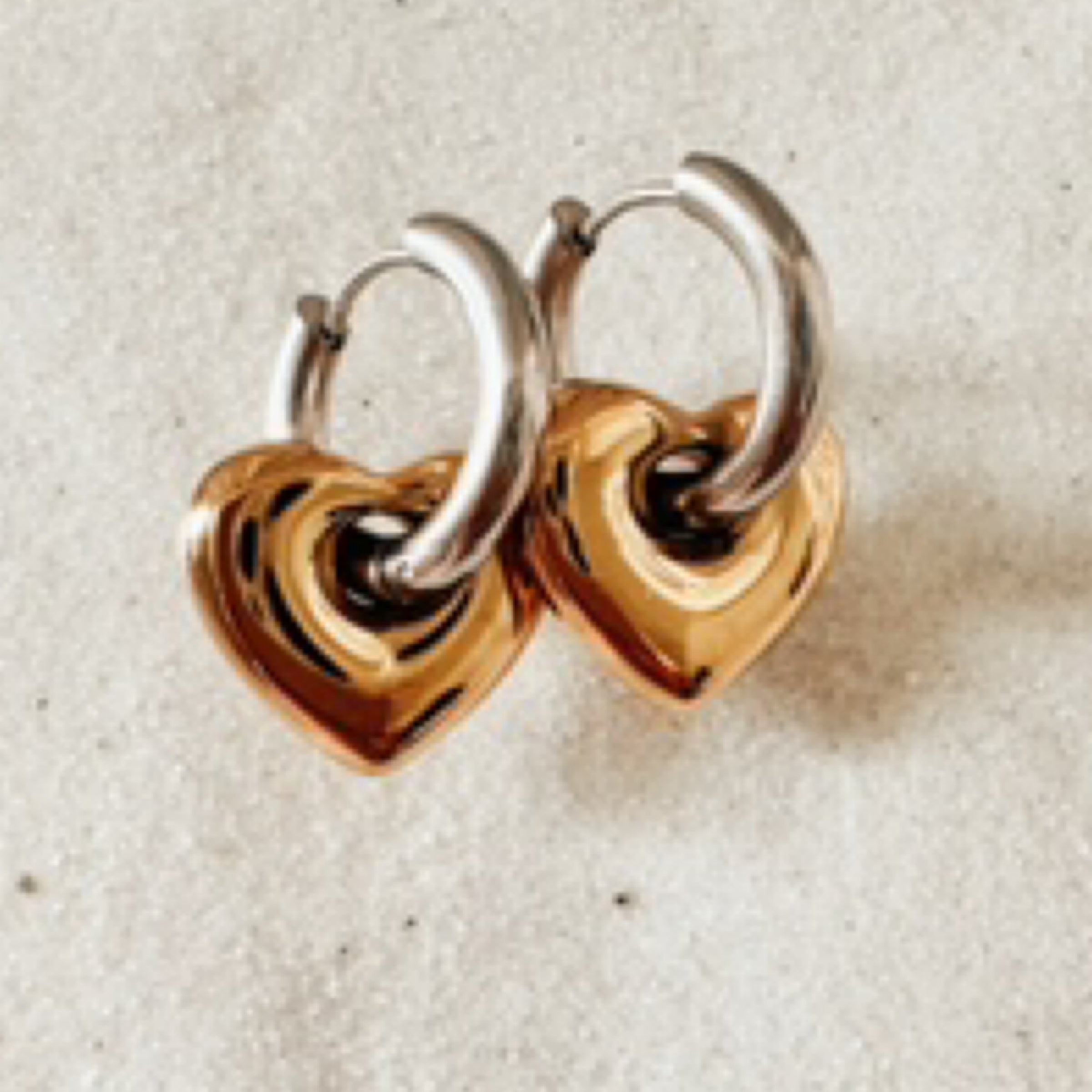 Silver huggies with large gold heart dangles. Pictured against white sand.