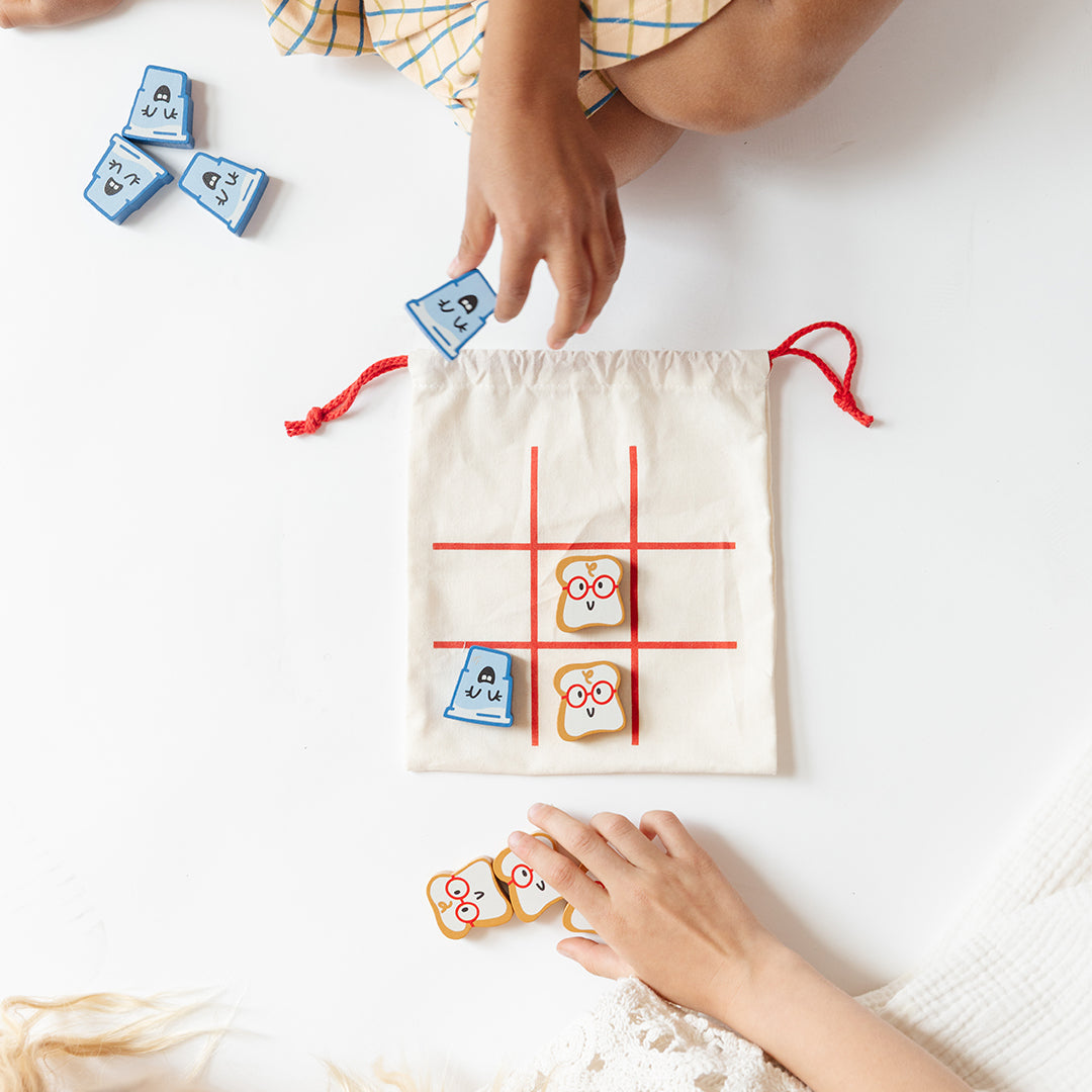 A blue and orange Tic Tac Toe game is shown with wooden game pieces featuring playful illustrated designs. The pieces are partially arranged on the game board beside an open cream-colored drawstring pouch with red cords.