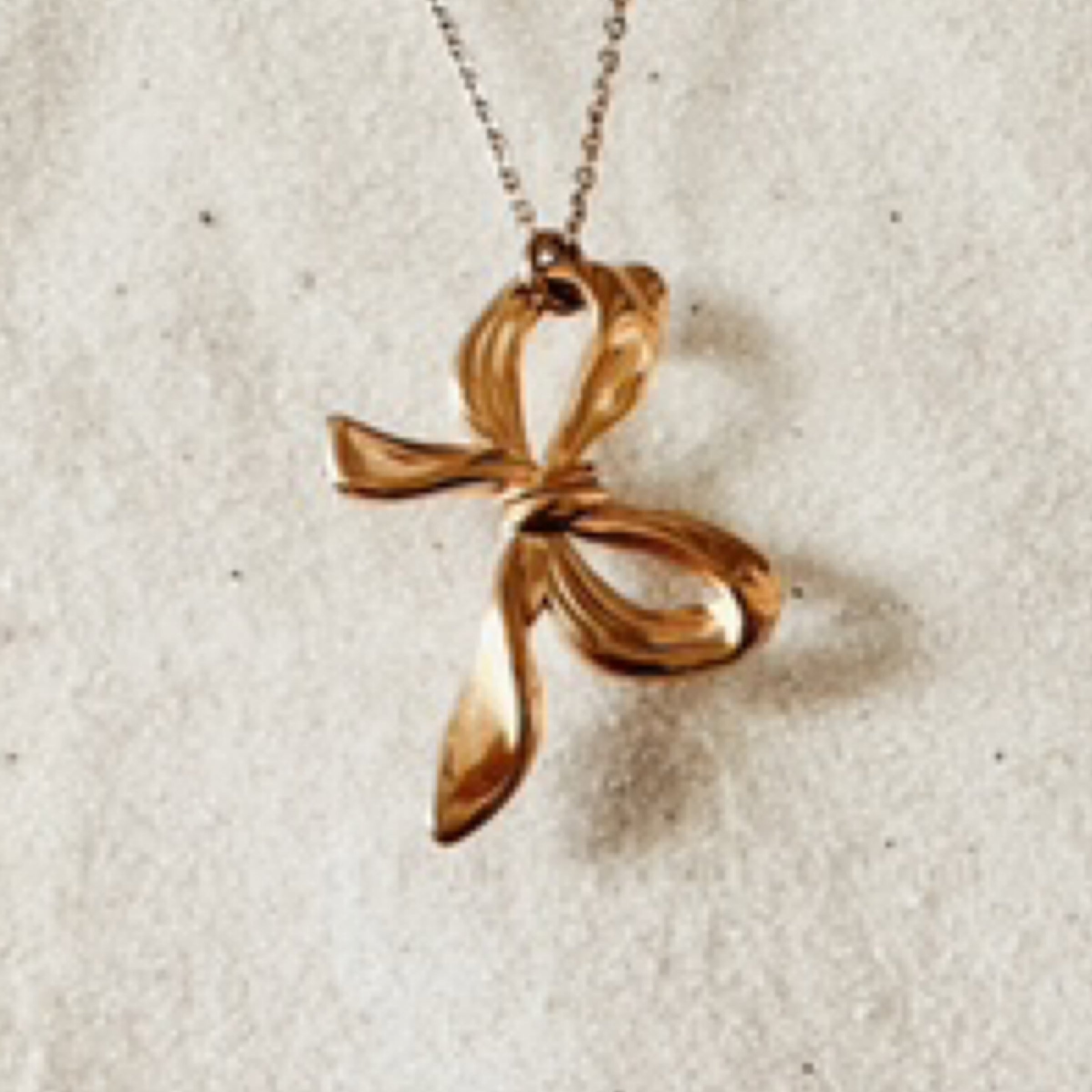 Delicate gold chain with a large life like gold bow charm hanging on a tilt. Pictured against white sand.