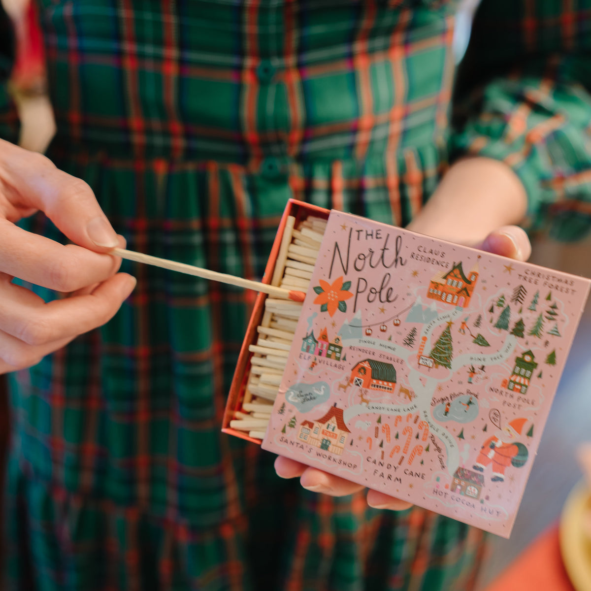 A person in a green plaid dress holds an open decorative matchbox labeled 'The North Pole' while lighting a match with a wooden stick. The matchbox has a pink illustrated cover with whimsical winter and travel-themed drawings.