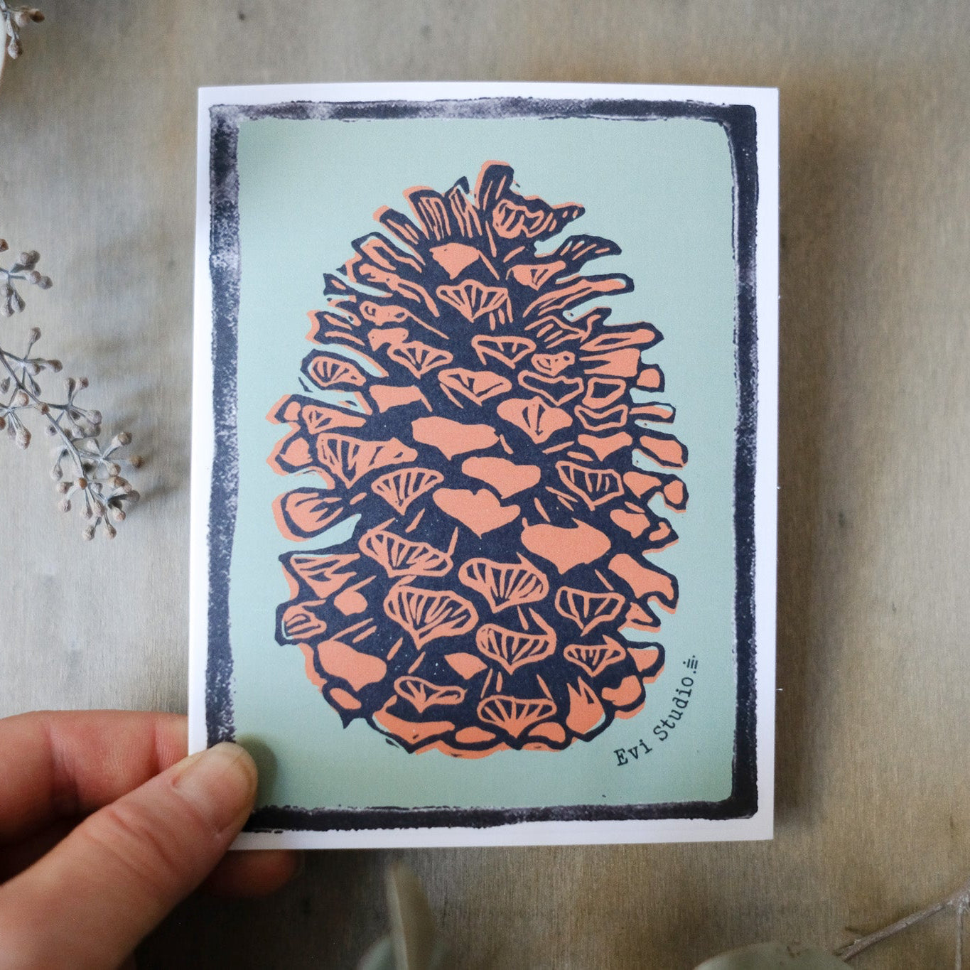 Rustic block print of a pine cone in warm brown with classic black ink detailing against a light sage green. Pictured against a wood board in a woman's hand.