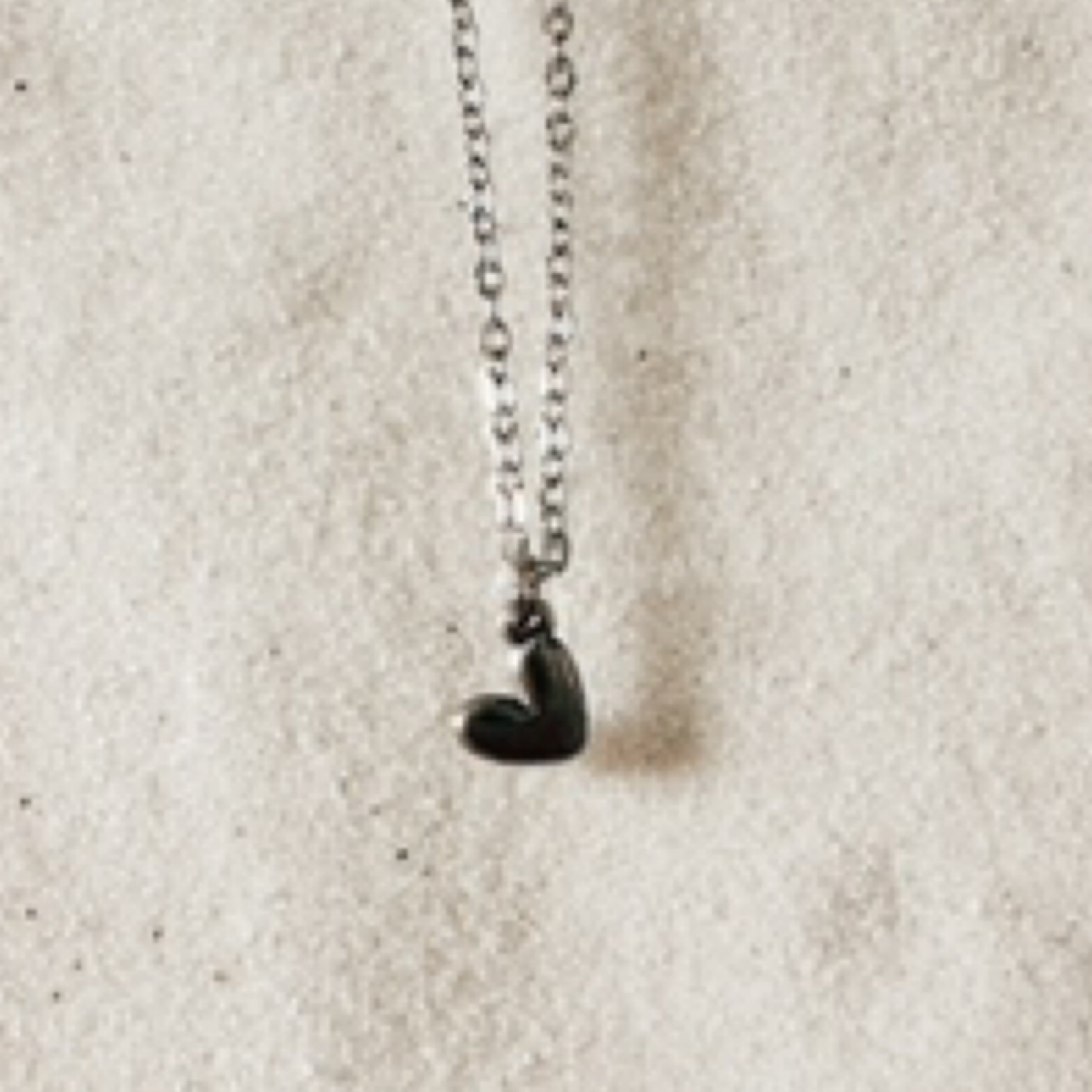 Delicate silver chain with a small flat heart pendant hanging on a tilt. Pictured against white sand.