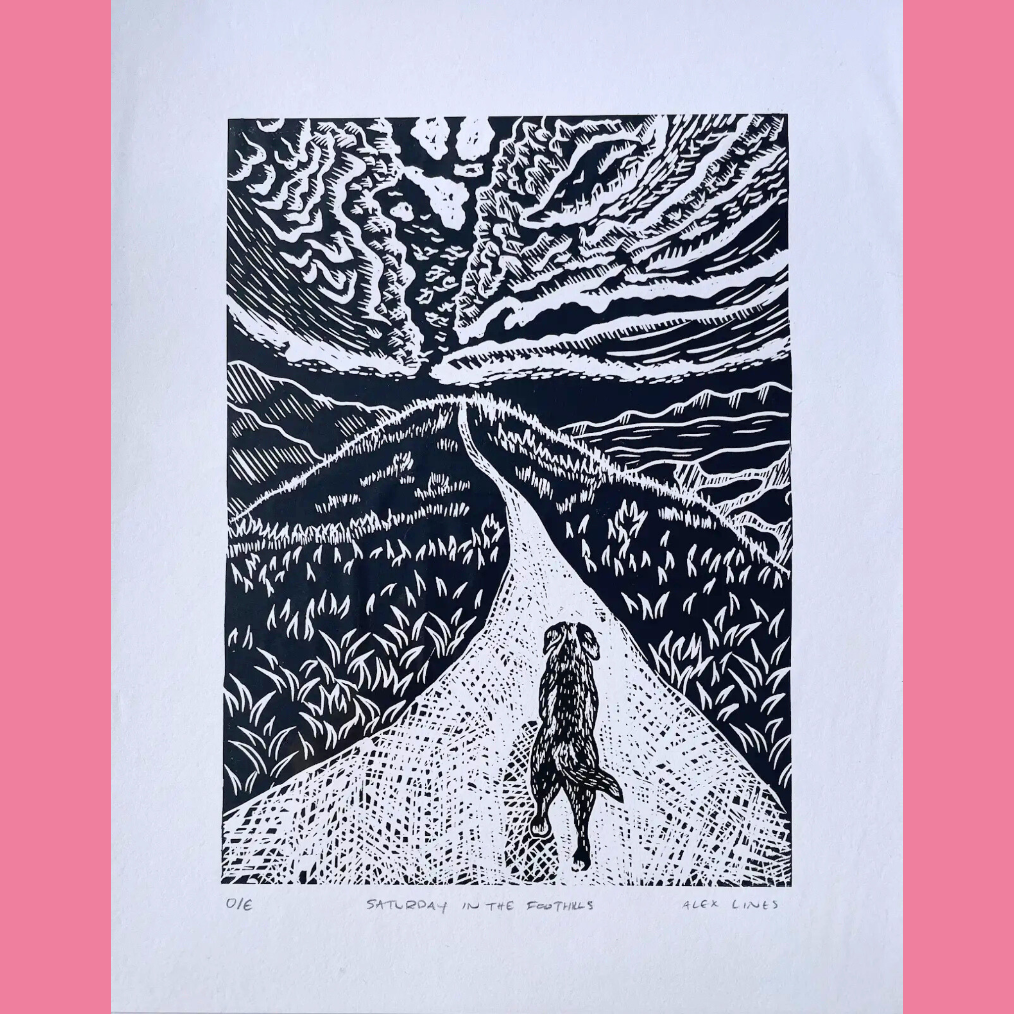 A black-and-white linocut print shows a dog walking away down a winding path through grassy foothills, viewed from behind as the trail stretches toward distant mountains under a dramatic, swirling sky filled with textured clouds and radiating lines. The artwork is printed on white paper with the title "SATURDAY IN THE FOOTHILLS," the number "016," and the signature "ALEX LINES" written below, surrounded by a bright pink border.
