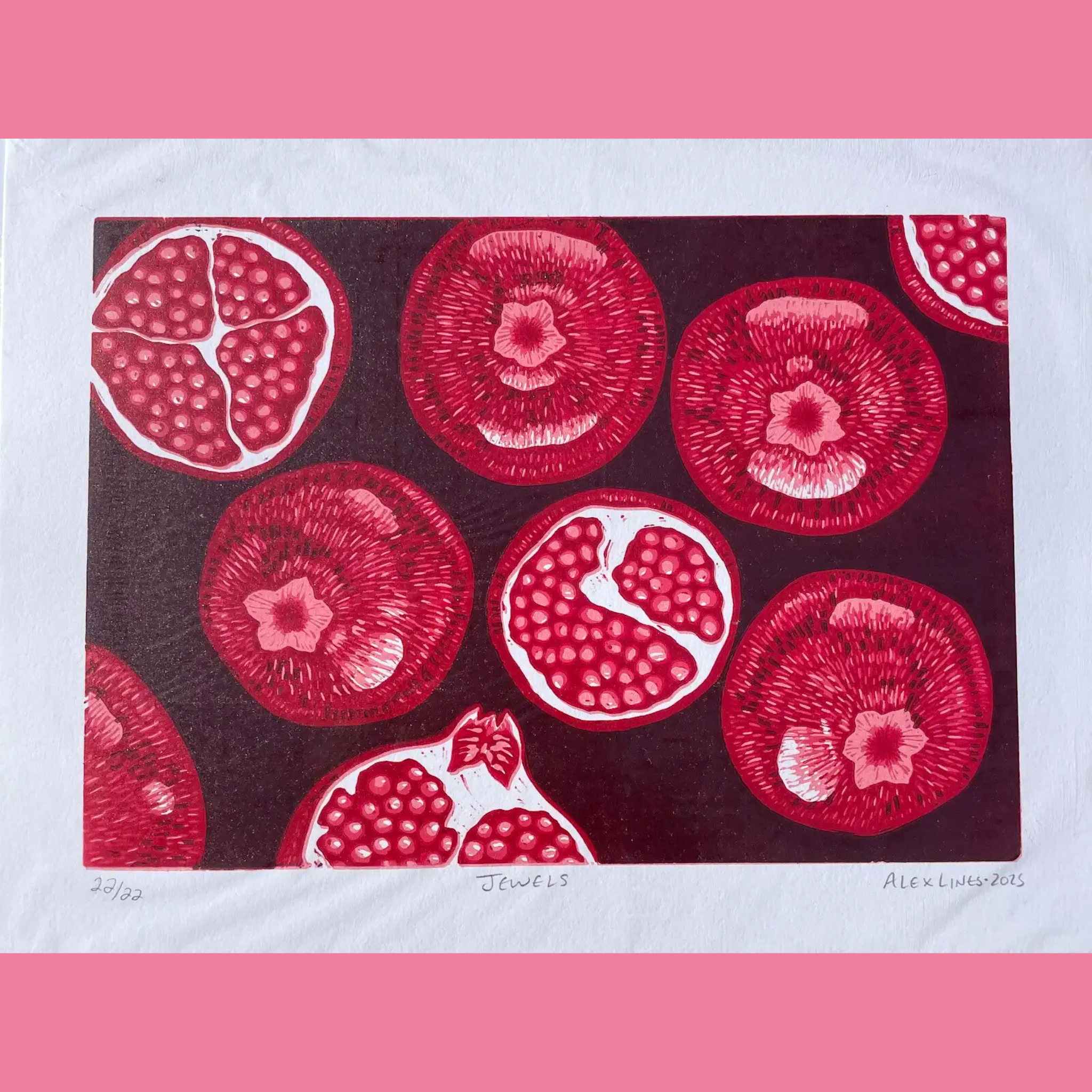 A vibrant linocut print features multiple cross-sections of pomegranates in rich shades of red and pink, arranged in a repeating pattern across a dark maroon background, with each halved fruit revealing clusters of juicy seeds and delicate white membranes, some showing a central star-shaped flower-like core. The artwork is printed on white paper with the edition number "20/22," title "JEWELS," and signature "ALEX LINES • 2025" handwritten below, surrounded by a bright pink border.