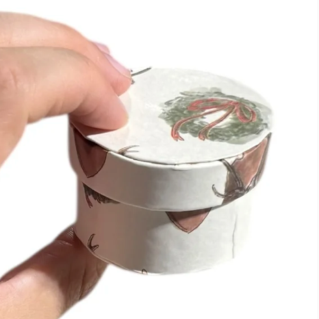 Hand holding a small round container with a decorative lid on a white background