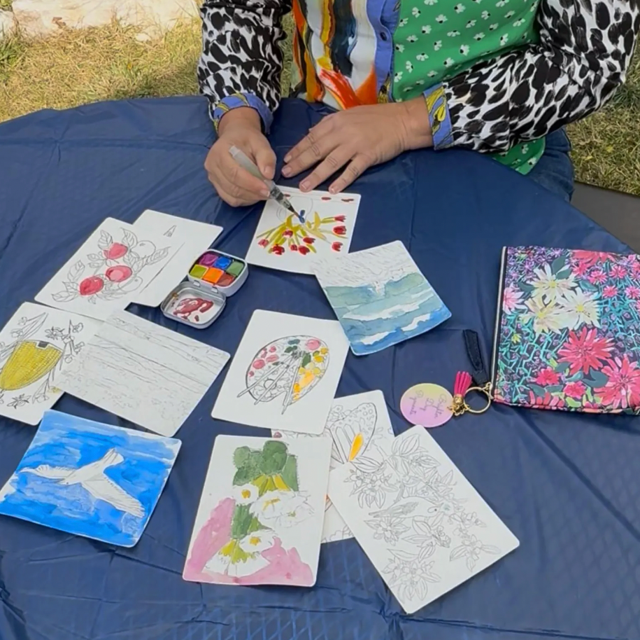 Person painting on paper with a watercolor set on a blue tablecloth outdoors.