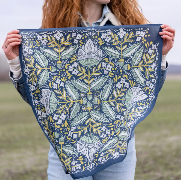 A person outdoors holds up a square scarf or bandana with an intricate botanical pattern in navy blue, sage green, white, and mustard yellow.
