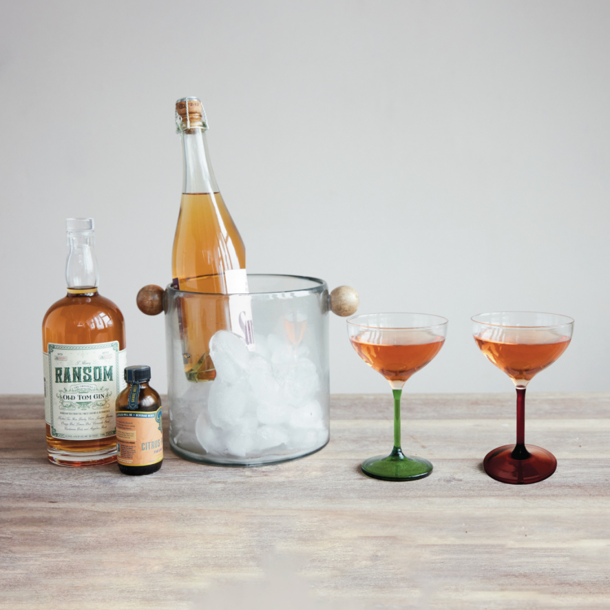 A styled tabletop scene with a clear glass bottle filled with amber liquid, a glass ice bucket with wooden handles and ice, two coupe glasses filled with pinkish-orange drink, and two bottles on a light wood surface against a pale gray background.