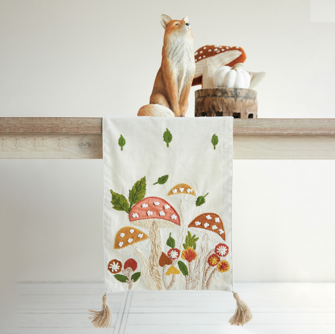 A cream fabric table runner with embroidered mushrooms and leaves, hanging from a wooden shelf. A small fox figurine and a basket of pumpkins sit on the shelf above.