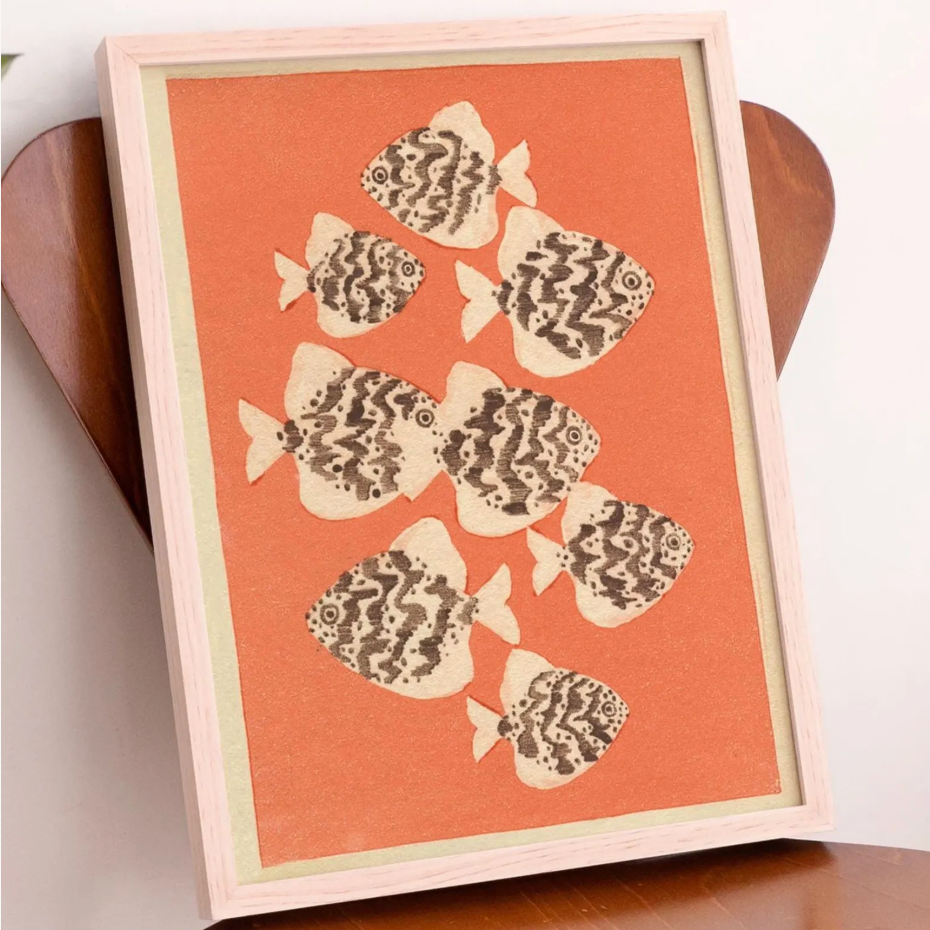 Framed wall art featuring seven stylized fish cutouts with dark brown wavy patterns on a warm orange background, set in a light natural wood frame and leaning against a wooden chair.