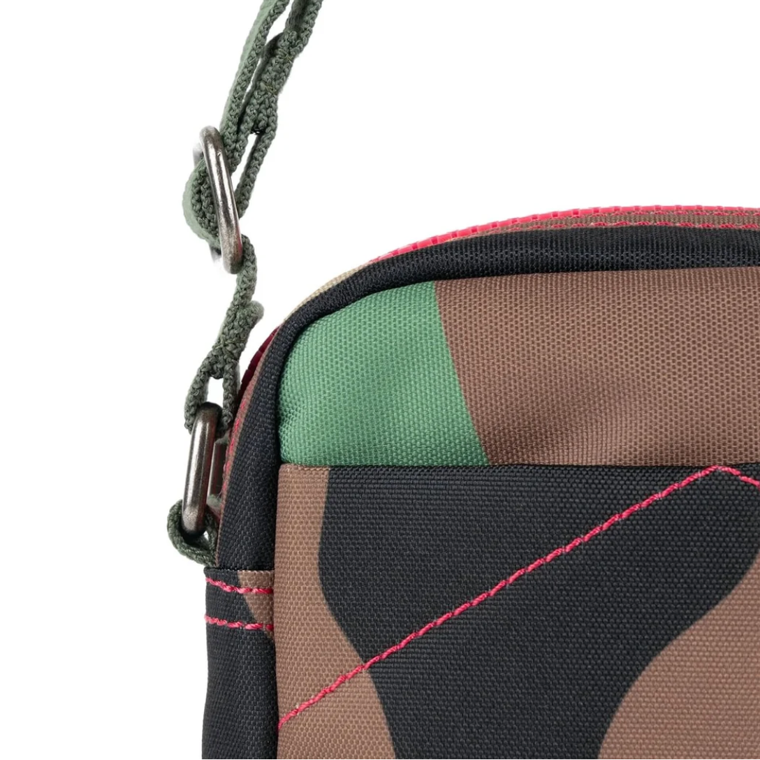 Close-up of a crossbody bag featuring a green, brown, and black camouflage pattern, pink contrast stitching, and a green woven strap with metal hardware, set against a plain white background.