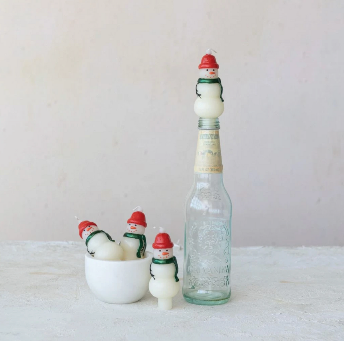 Three snowman-shaped candles with red hats and green scarves are displayed on a light surface against a soft beige background. One candle is standing upright in a clear glass bottle, while the other two are grouped in a small white bowl.