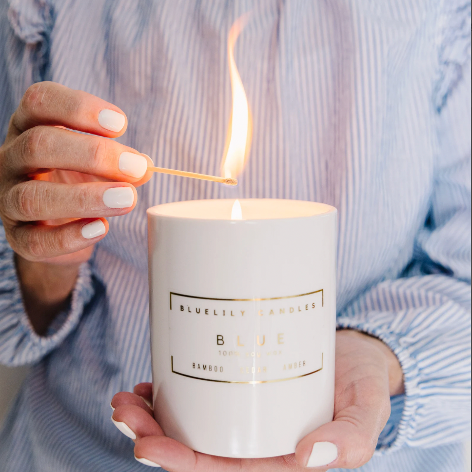 A person in a blue and white striped shirt holds a lit white candle labeled 'Blue Bamboo + Sandalwood' while lighting it with a wooden match. The candle is shown in a close-up lifestyle shot.