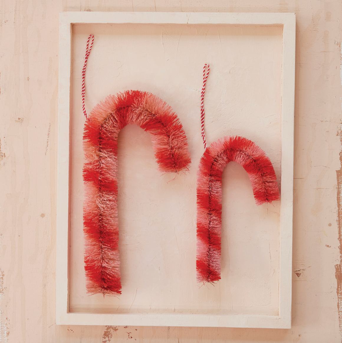 Two red and white fuzzy candy cane ornaments hanging from red-and-white twine inside a white wooden frame, displayed against a light beige wall.