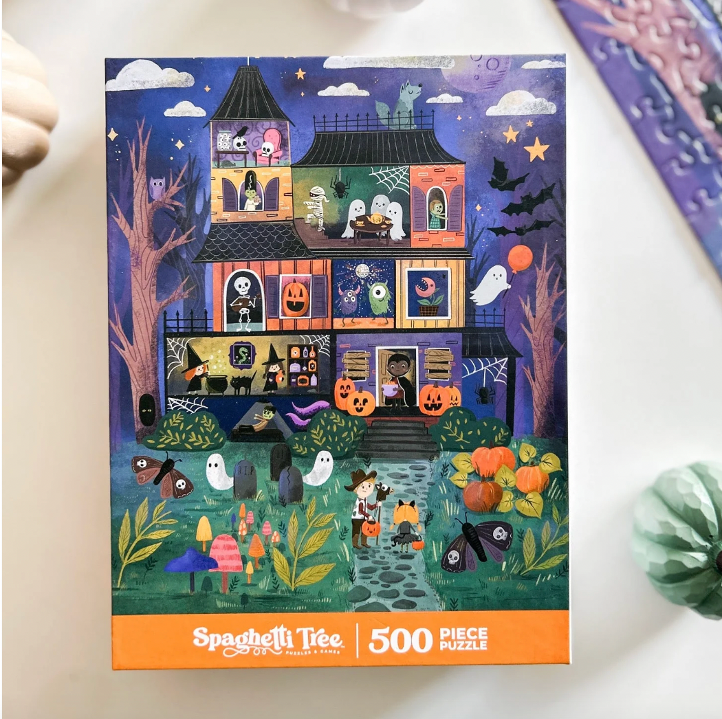 Spaghetti Tree 500-piece jigsaw puzzle box featuring a colorful cartoon illustration of a haunted house at Halloween, photographed on a light surface with small decorative props nearby.