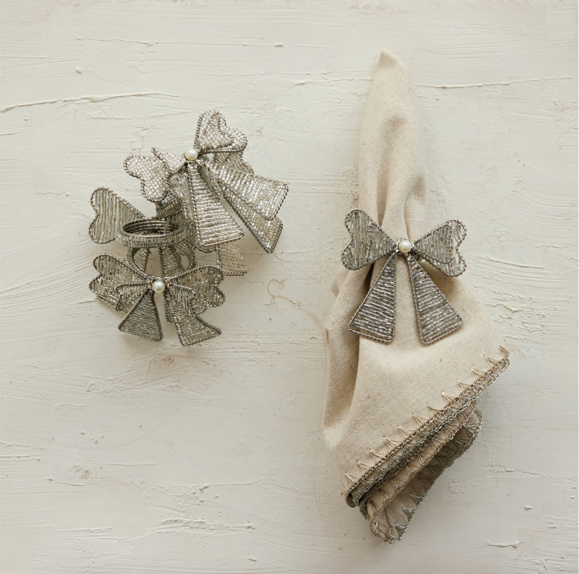 Two metallic silver bow-shaped napkin rings with small pearl-like centers, shown beside a folded cream linen napkin on a textured off-white background.