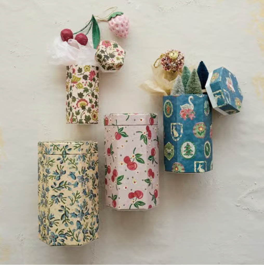 Four decorative cylindrical gift boxes with floral and fruit prints are arranged on a textured off-white wall, each holding small wrapped presents and decorative items.