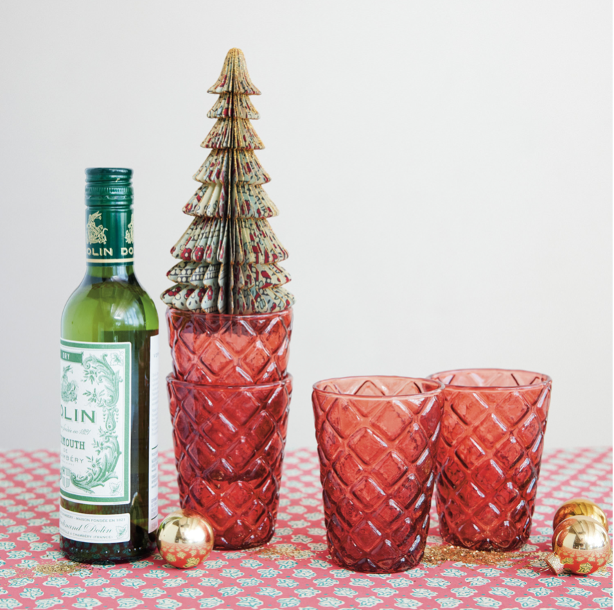 A styled tabletop scene with a green bottle on the left, a red faceted glass vase holding a small decorative tree in the center, and two matching red faceted drinking glasses on the right, all arranged on a patterned red-and-white cloth with small gold ornaments against a plain light background.